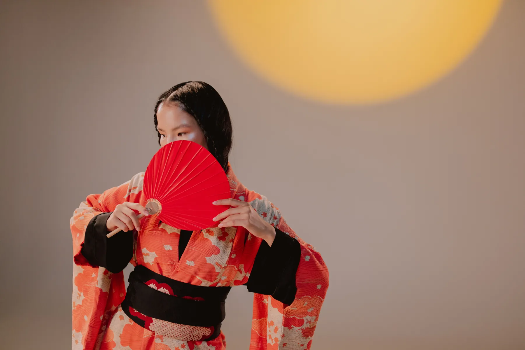A woman in an orange and black kimono holds a red folding fan in front of her face in warm studio light